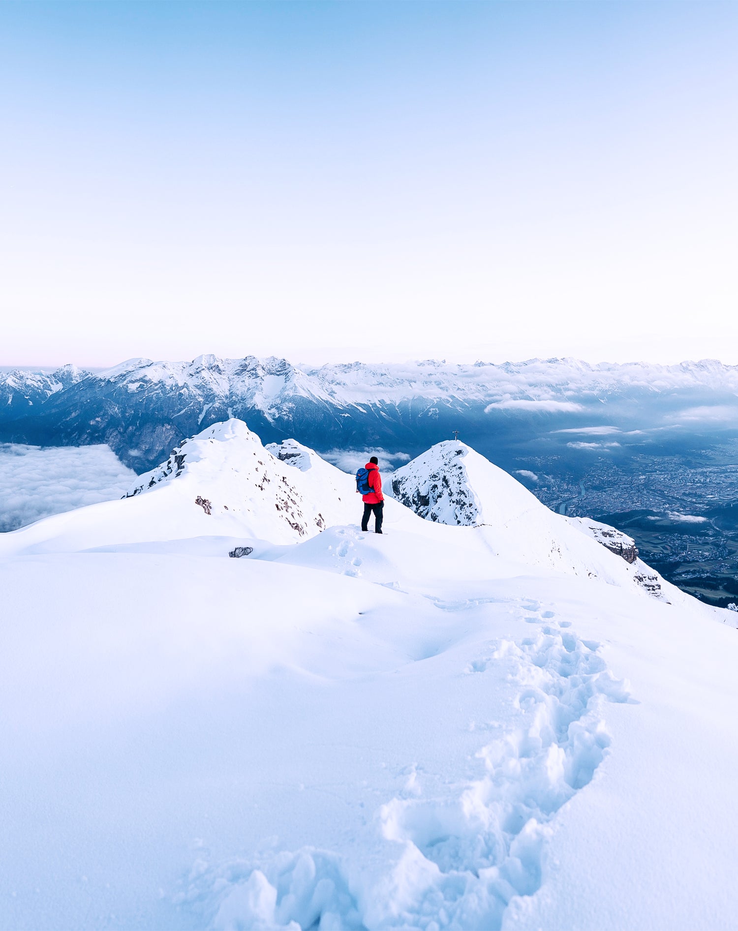 Tracks in the snow leading to a person on a mountain top with a red jacket contrasting to an epic blue horizon with a mountain range in the distance.