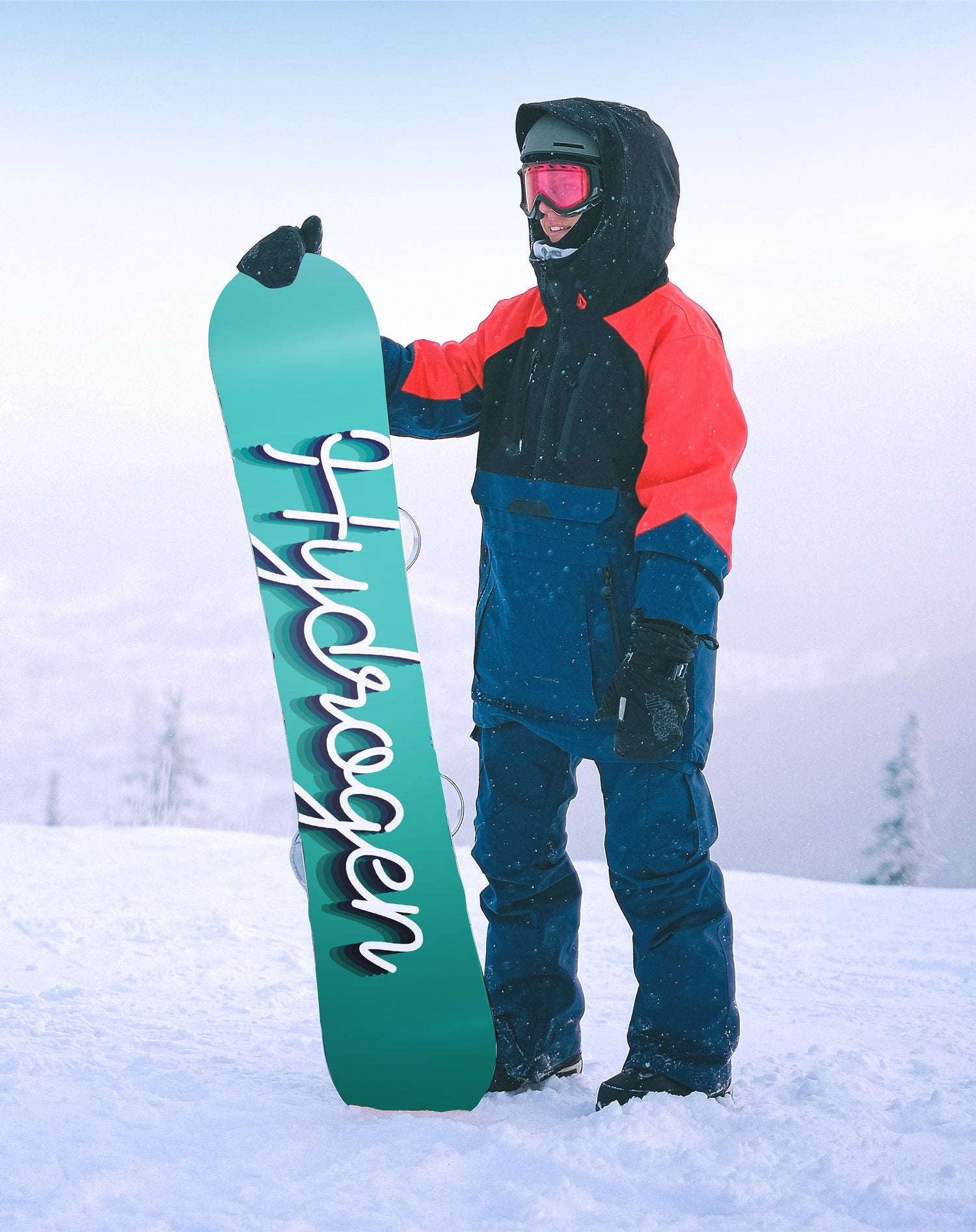 A snowboarder standing on a mountain top in choppy snow, shows off the back of his snowboard which reads Hydrogen in a cursive script.